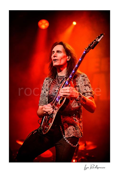 Vertical color image of Steve Vai playing guitar under vivid red stage lighting with glowing fret markers and light stage smoke