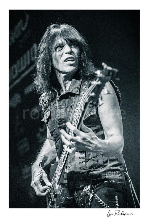 Vertical black and white image of Rudy Sarzo performing live with bass guitar in a close-up stage portrait