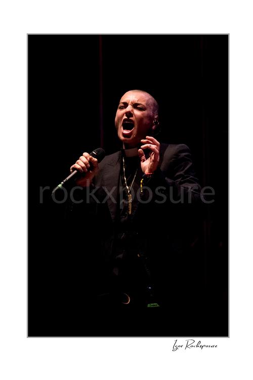 Vertical color image of Sinéad O’Connor singing into a microphone in a dramatic low-light stage portrait
