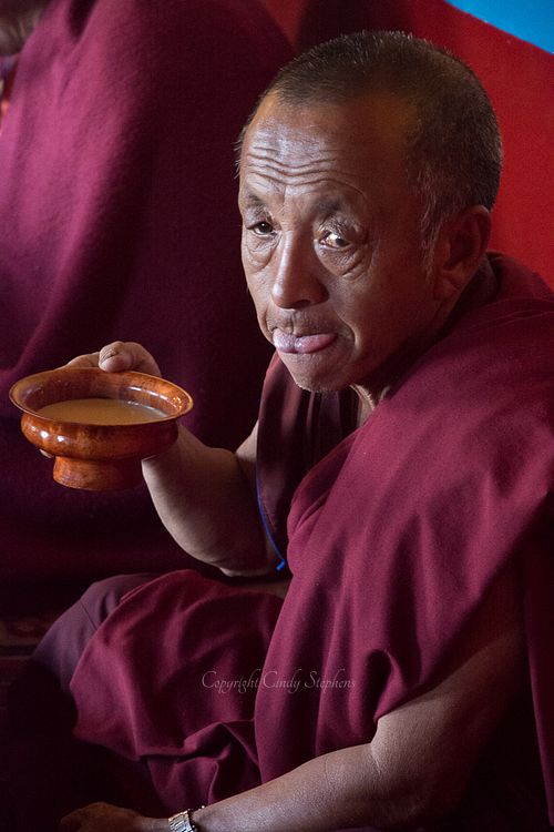 A Nepalese monk dressed in traditional crimson robes sits peacefully, sipping from a wooden bowl, embodying the tranquility of Buddhist monastic life.
