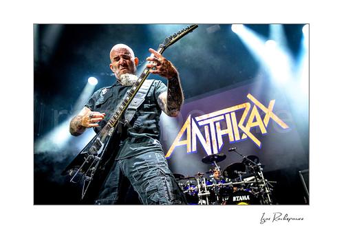 Horizontal color image of Scott Ian of Anthrax performing live on stage, playing guitar in front of the Anthrax stage backdrop under bright concert lighting