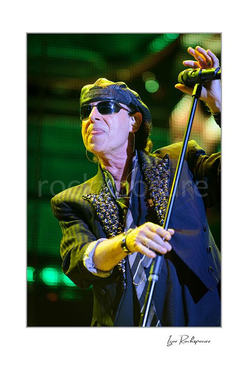 Vertical color image of Klaus Meine of Scorpions performing live in sunglasses and leather cap with microphone stand against green stage lights