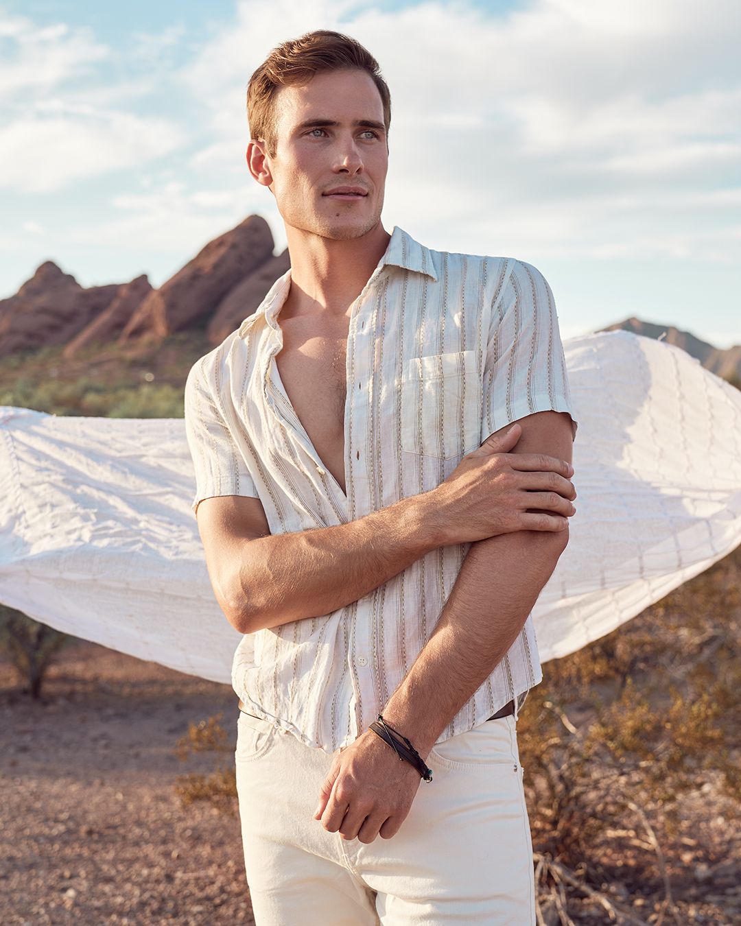 Male model posing outdoors in Phoenix, AZ during an editorial fashion shoot wearing a white short sleeve button down shirt, white pants, and a sheer white sheet blowing behind him in the breeze