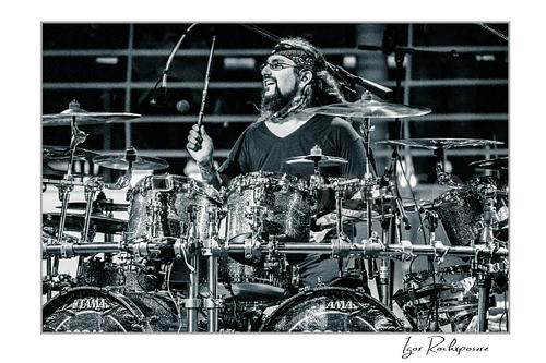 Horizontal black and white image of Mike Portnoy smiling at the drum kit during a live performance
