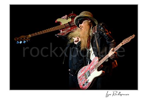 Horizontal color image of Billy Gibbons and Dusty Hill of ZZ Top performing live on stage, with Gibbons playing a red Telecaster while Hill lifts his red bass behind him against a dark background