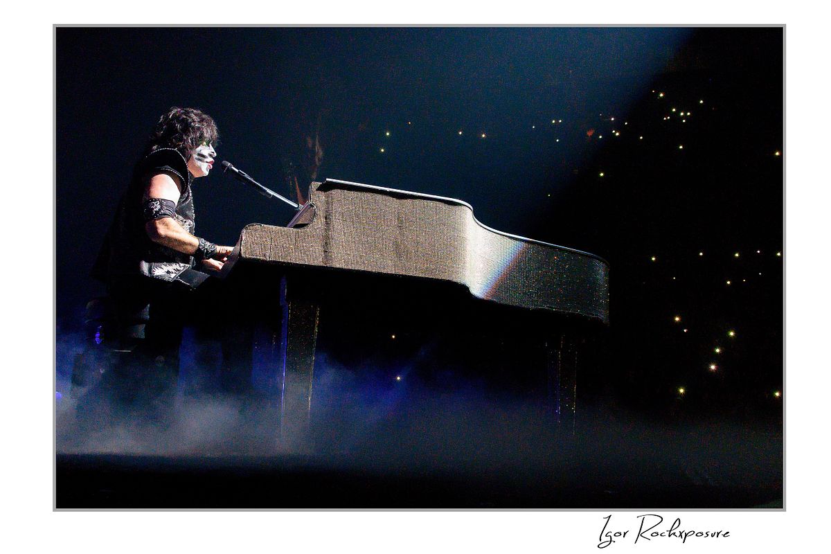 Horizontal color photograph of Eric Singer in full KISS makeup and costume seated at a glittering grand piano while singing under soft concert lighting with scattered lights visible in the background