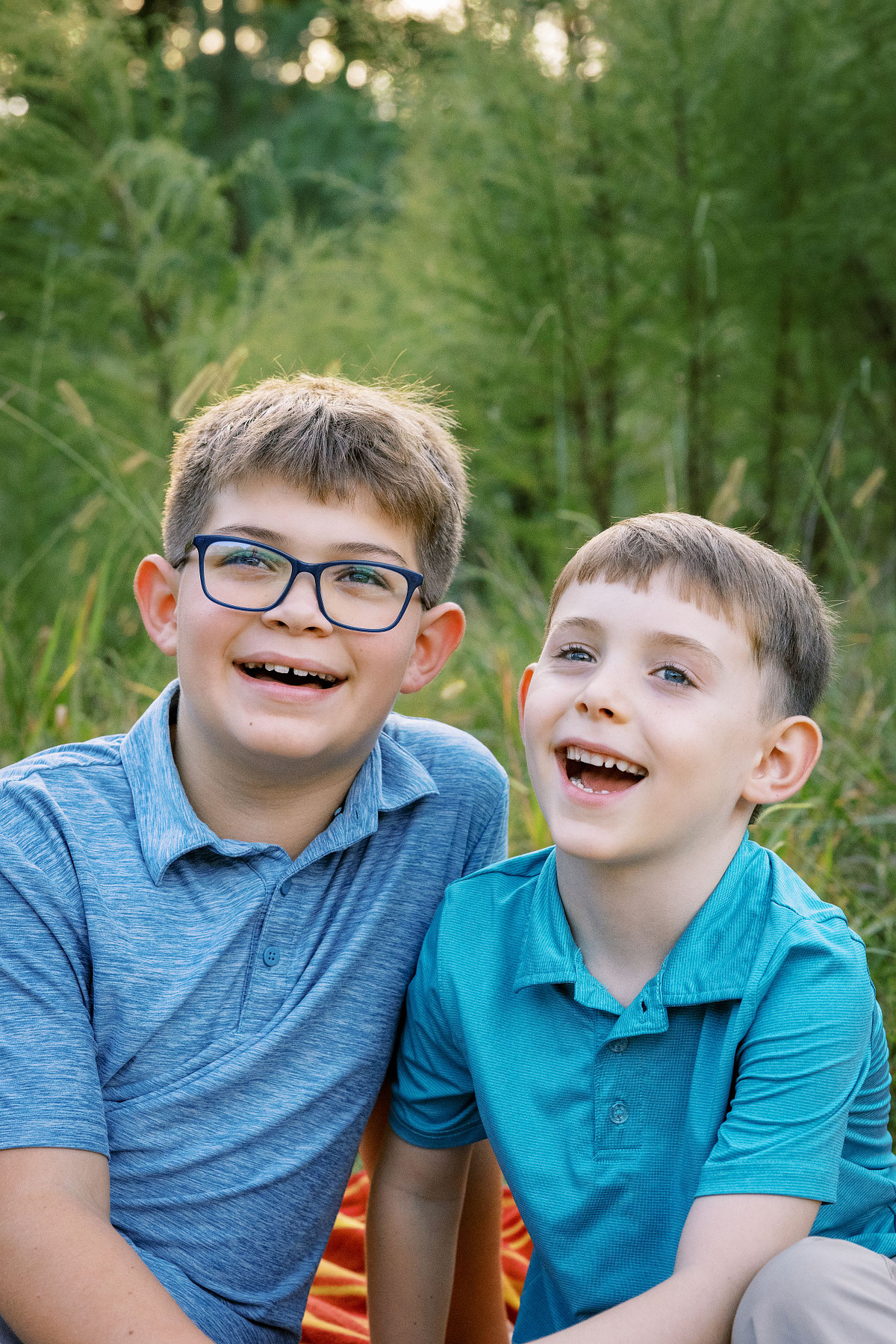 Two smiling young brothers in coordinating blue polo shirts enjoy a summer family photoshoot in North Carolina, sitting on a blanket in a grassy field during golden hour.