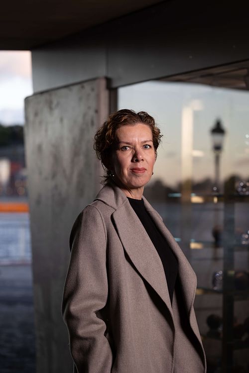 Halvkroppsporträtt av kvinna i kappa i skymning vid glasfasad med reflektioner, personal branding i Stockholm.