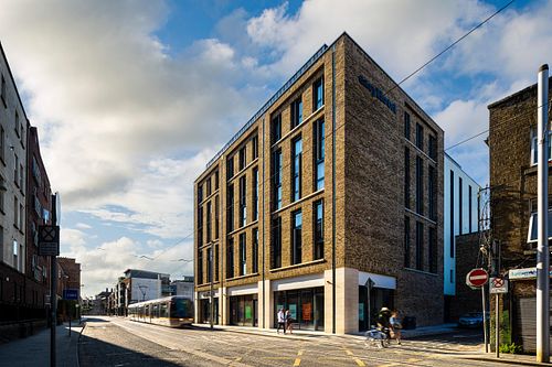 Street-level view of the Easy Hotel Dublin, showing its modern brick facade and surrounding urban environment. A tram is passing on the street, and pedestrians are visible. The 'easyHotel Dublin City Centre' sign is on the building. A Lycamobile shop is on the right commercial side of the building.