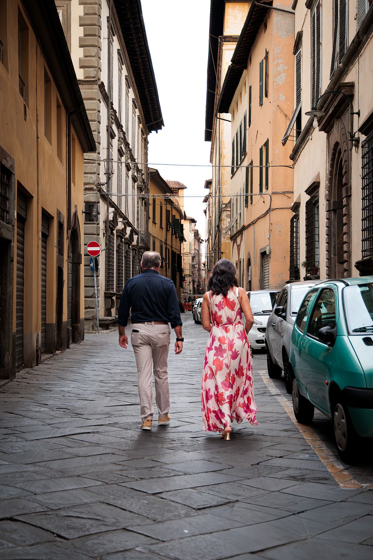 Couple walking away down a narrow cobblestone street in Lucca, Italy, with warm yellow buildings on the left, parked cars including a light green one on the right, the woman in a long pink floral dress and the man in a dark blue shirt and beige pants, under soft overcast light creating a calm, romantic atmosphere.