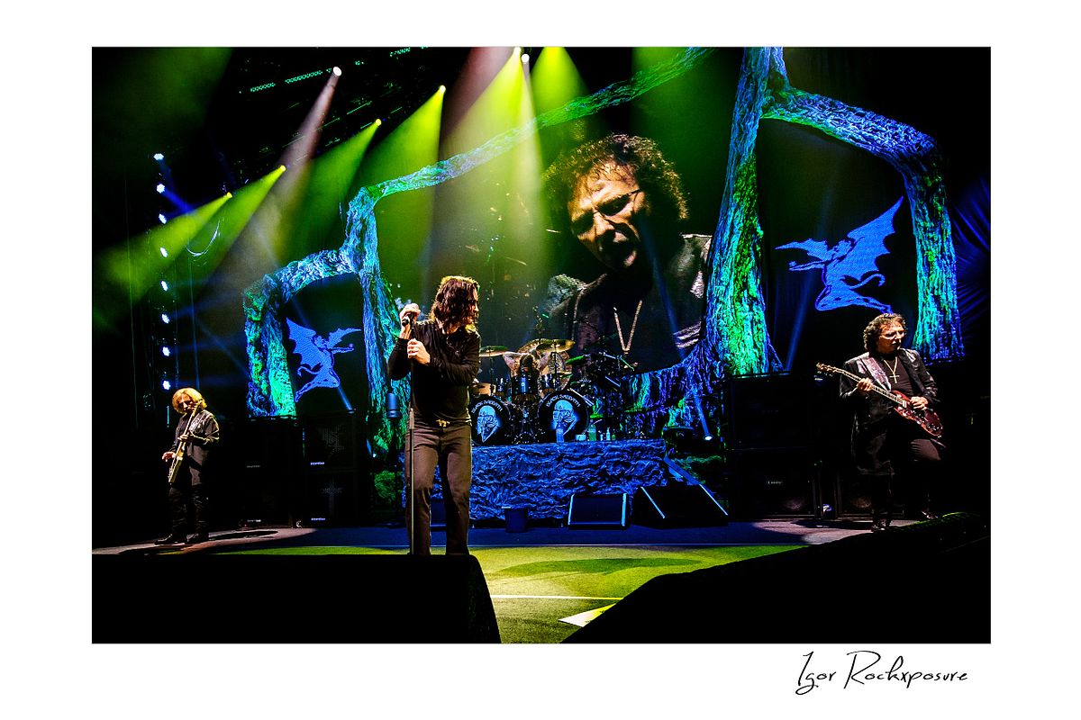 Black Sabbath performing on stage with all four band members under dramatic green and yellow concert lighting with a giant screen showing Tony Iommi behind them