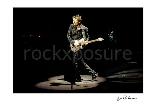 Horizontal color image of Matt Bellamy of Muse playing guitar alone in a spotlight on a dark stage