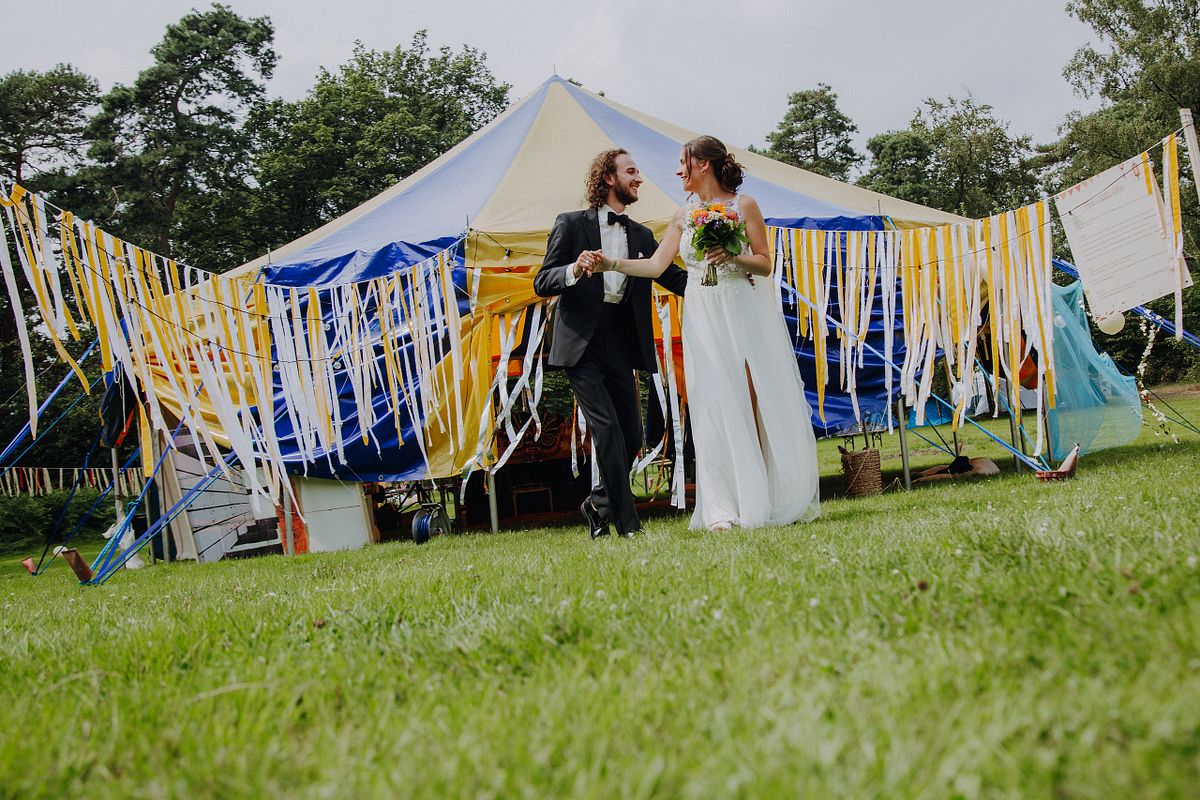 Alternativ gestyltes Brautpaar posiert vor einem bunten Festzelt oder Tipizelt, unkonventionelle Festival-Hochzeit in NRW und Umgebung.