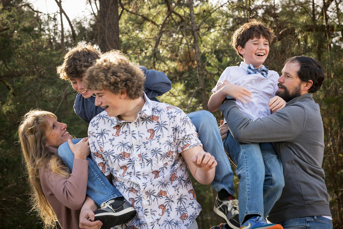A candid and playful photo of parents lifting and playing with their three laughing sons during an outdoor family photoshoot in a wooded park.