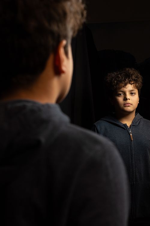 Pojke i mörk hoodie ser på sin spegelbild i lågmält ljus, svart bakgrund