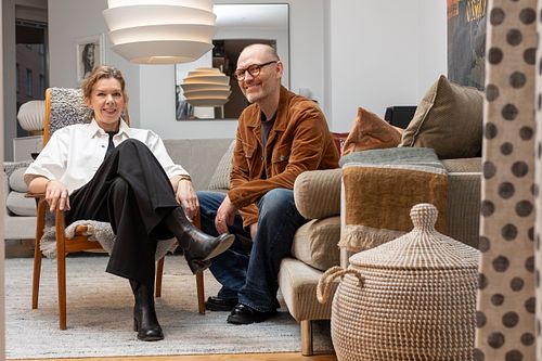 Två personer sitter i ett skandinaviskt vardagsrum med textilier och detaljer, fotograferat i redaktionell stil.