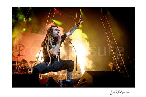 Horizontal color image of Randy Blythe of Lamb of God crouched on stage with long dreadlocks, handheld microphone, and a burst of fire behind him