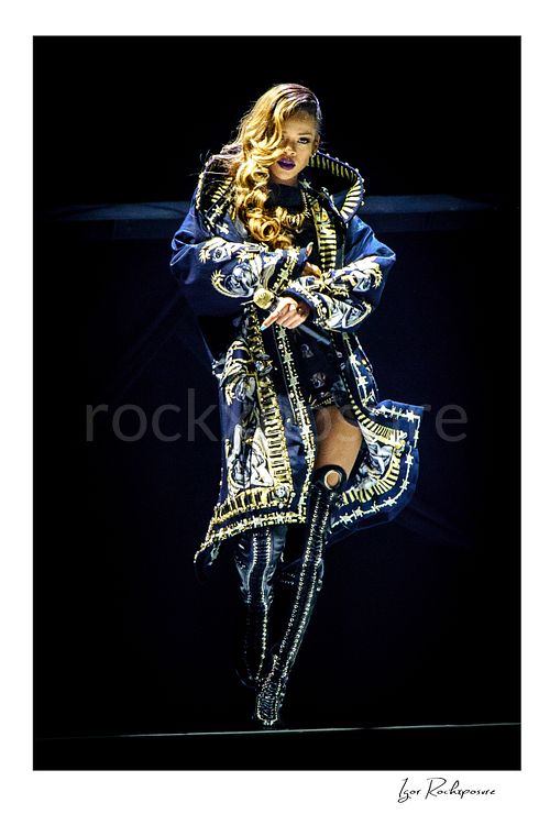 Vertical color image of Rihanna performing on stage in an ornate coat and thigh-high boots against a dark concert background