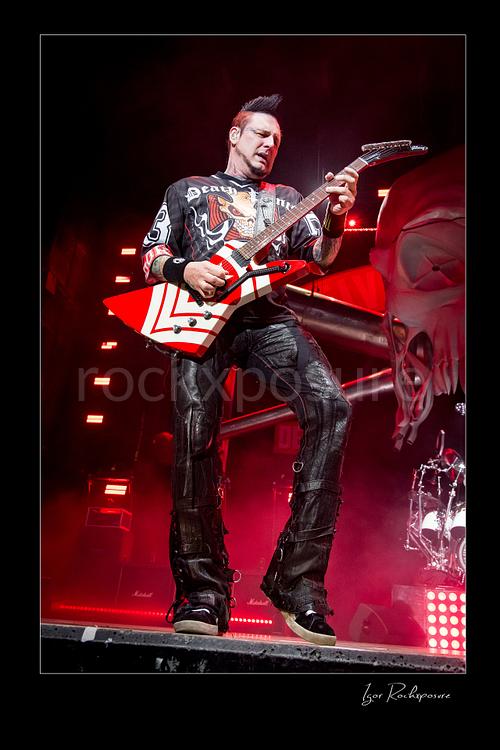 Vertical color image of Zoltan Bathory of Five Finger Death Punch playing guitar onstage under red concert light beside a giant skull stage prop