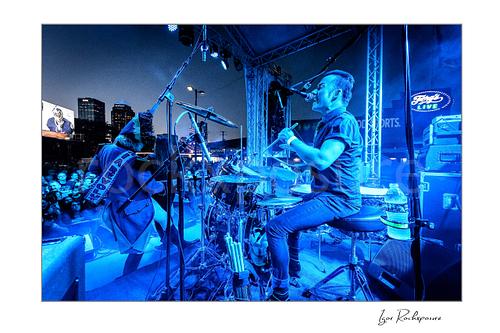Horizontal color image of Stig Pedersen and Laust Sonne performing on stage in blue concert light, with Laust Sonne singing at the drum kit and a crowd visible beyond the stage