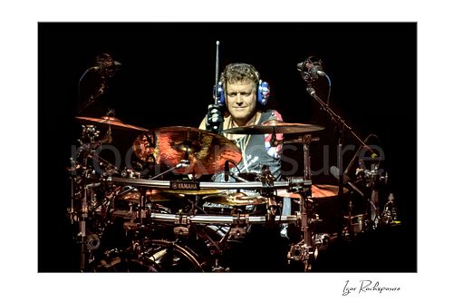 Horizontal color image of Rick Allen of Def Leppard performing live on stage behind his drum kit, wearing Union Jack headphones and holding a drumstick under concert lighting