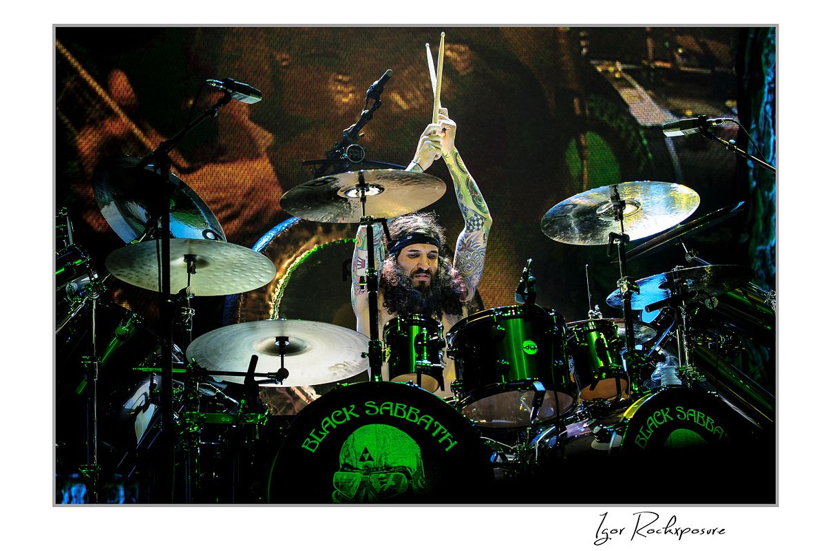 Tommy Clufetos of Black Sabbath raising drumsticks while playing drums on stage under green concert lighting