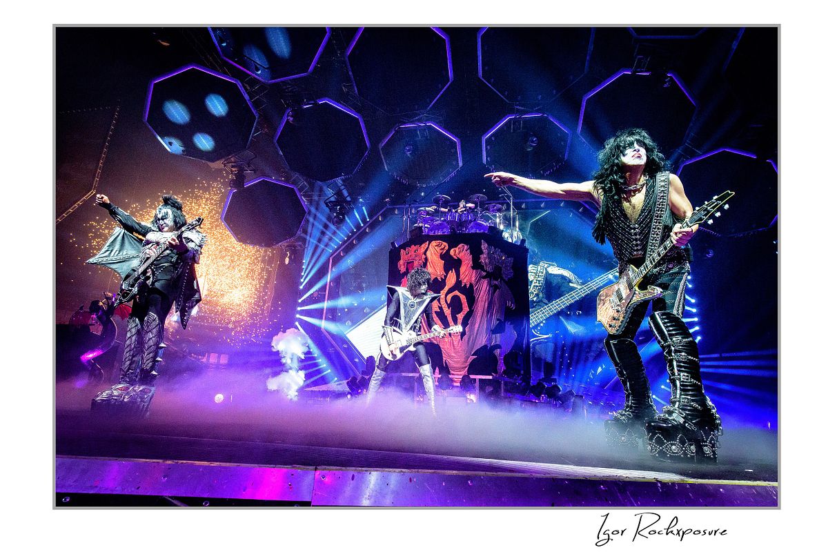 Horizontal color photograph of Gene Simmons, Paul Stanley, and Tommy Thayer performing onstage in full makeup and costumes surrounded by fog, bright lighting structures, and intense concert effects