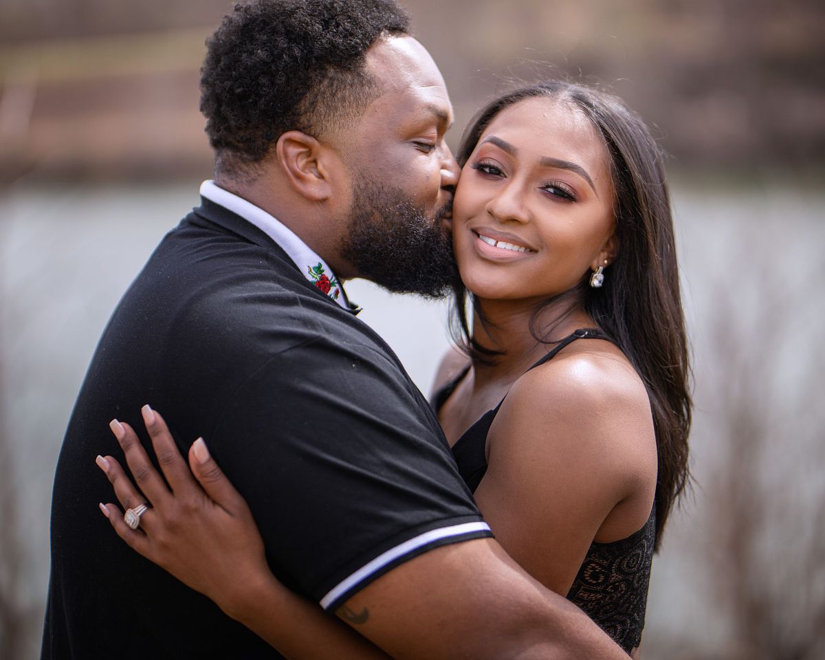 black couple embracing, the woman is smiling at the camera while the man is kissing her cheek.