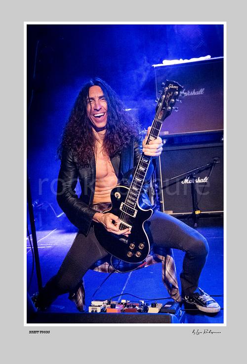 Vertical color image of Brent Woods playing guitar on stage with a wide smile, open jacket, and blue stage light