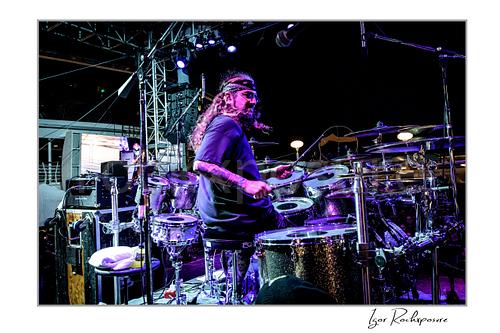 Horizontal color image of Mike Portnoy playing drums from the side under purple stage lighting on a nighttime outdoor stage