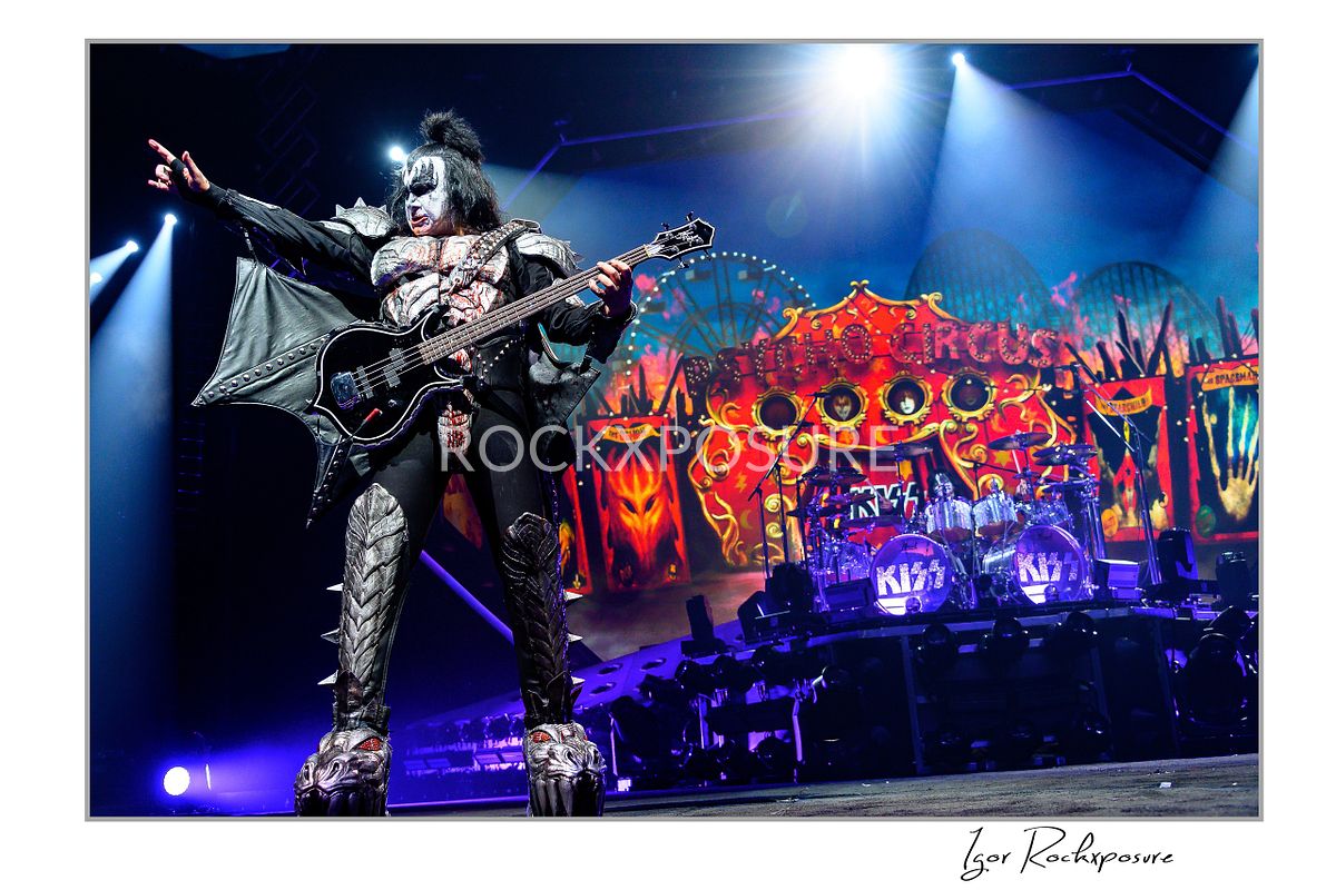 Gene Simmons of KISS performing in a wide power stance with bass guitar under bright white and blue stage lighting with fiery stage visuals behind him