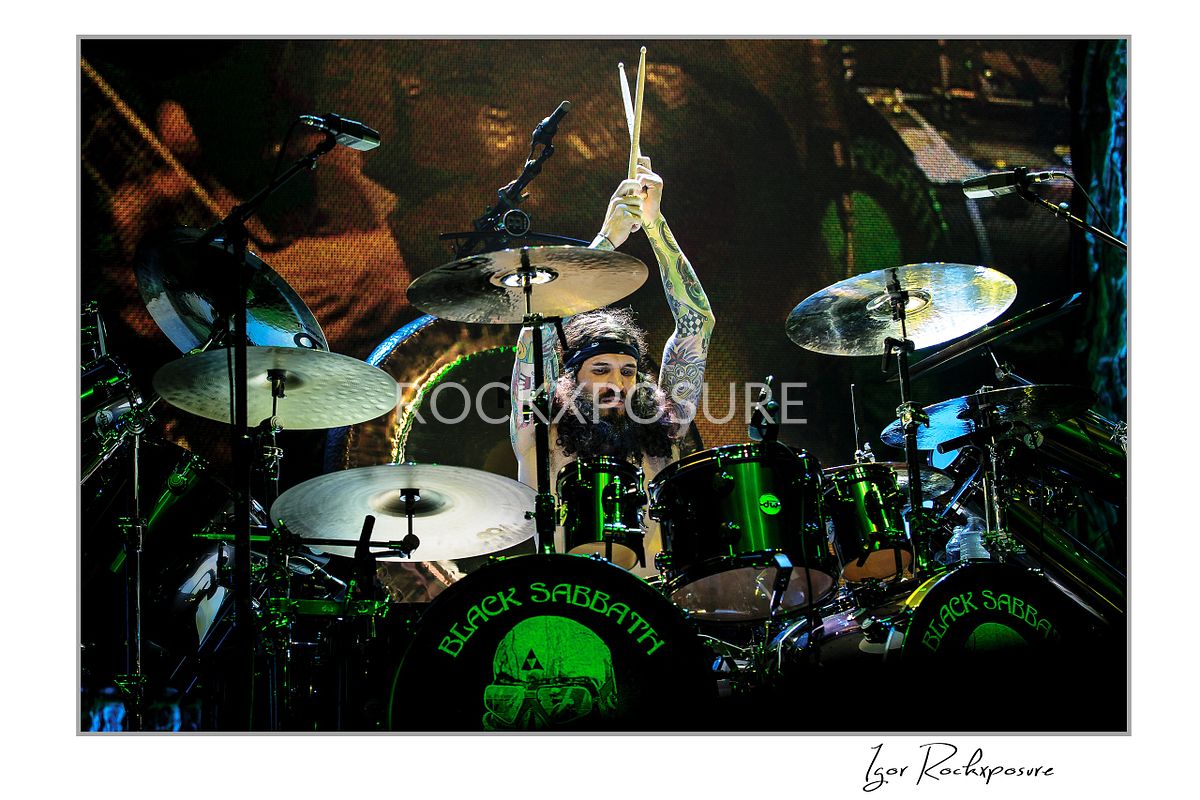 Tommy Clufetos of Black Sabbath raising drumsticks while playing drums on stage under green concert lighting