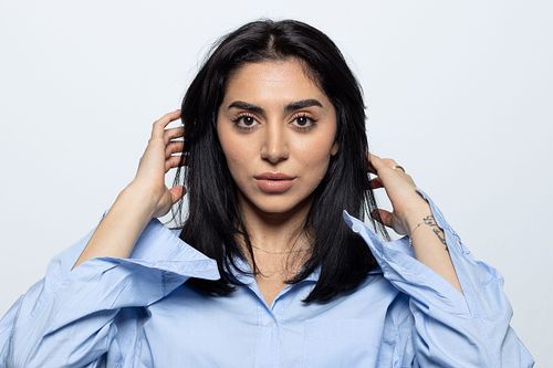 Kvinna i ljus blå skjorta ser in i kameran och håller händerna vid håret, vit bakgrund