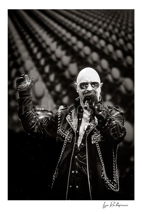 Vertical black and white image of Rob Halford of Judas Priest performing live on stage, singing into a microphone while wearing sunglasses and a studded leather coat