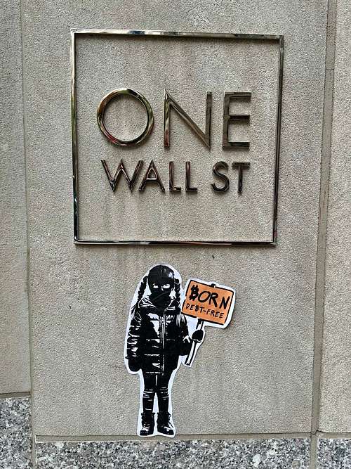 Street art sticker by Street Cyber in New York City, placed directly in front of the "ONE WALL ST" building sign. The sticker depicts a child figure in a black ski mask and jacket, holding an orange sign that reads "BORN DEBT-FREE." This powerful piece directly critiques traditional financial systems and advocates for individual financial freedom and sovereignty through Bitcoin, free from inflationary debt.