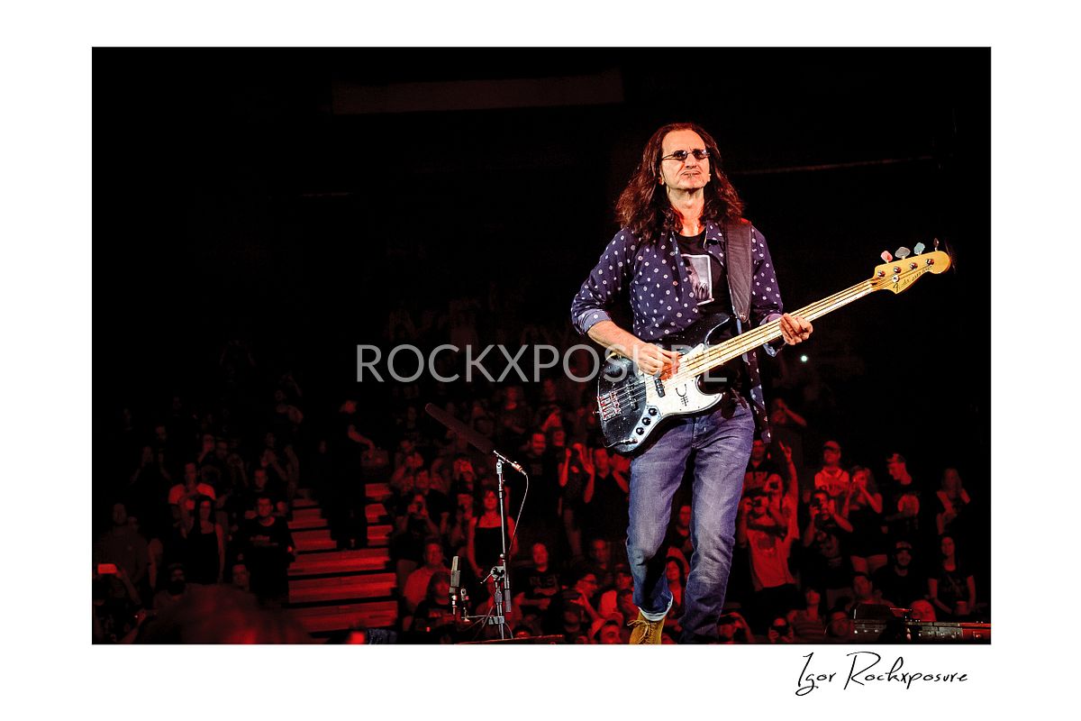 Horizontal concert photography of Geddy Lee playing bass guitar on stage with audience visible under stage lighting
