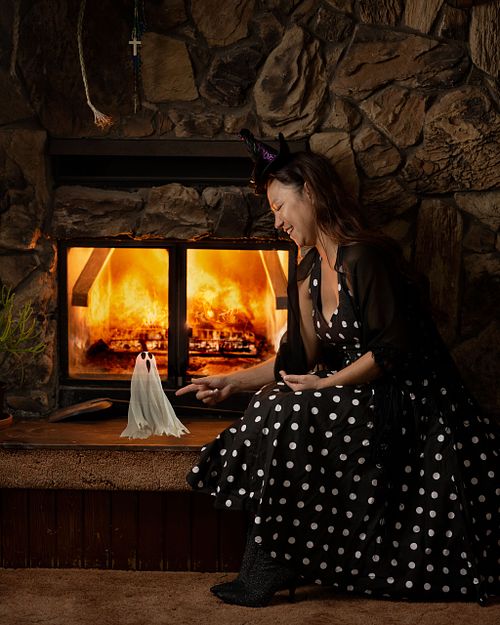 A fancy witch pokes her little ghost friend in front of a fireplace.