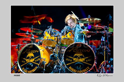 Horizontal color image of Rikki Rockett performing at the drum kit during a Poison concert, surrounded by cymbals with vivid red and blue stage light