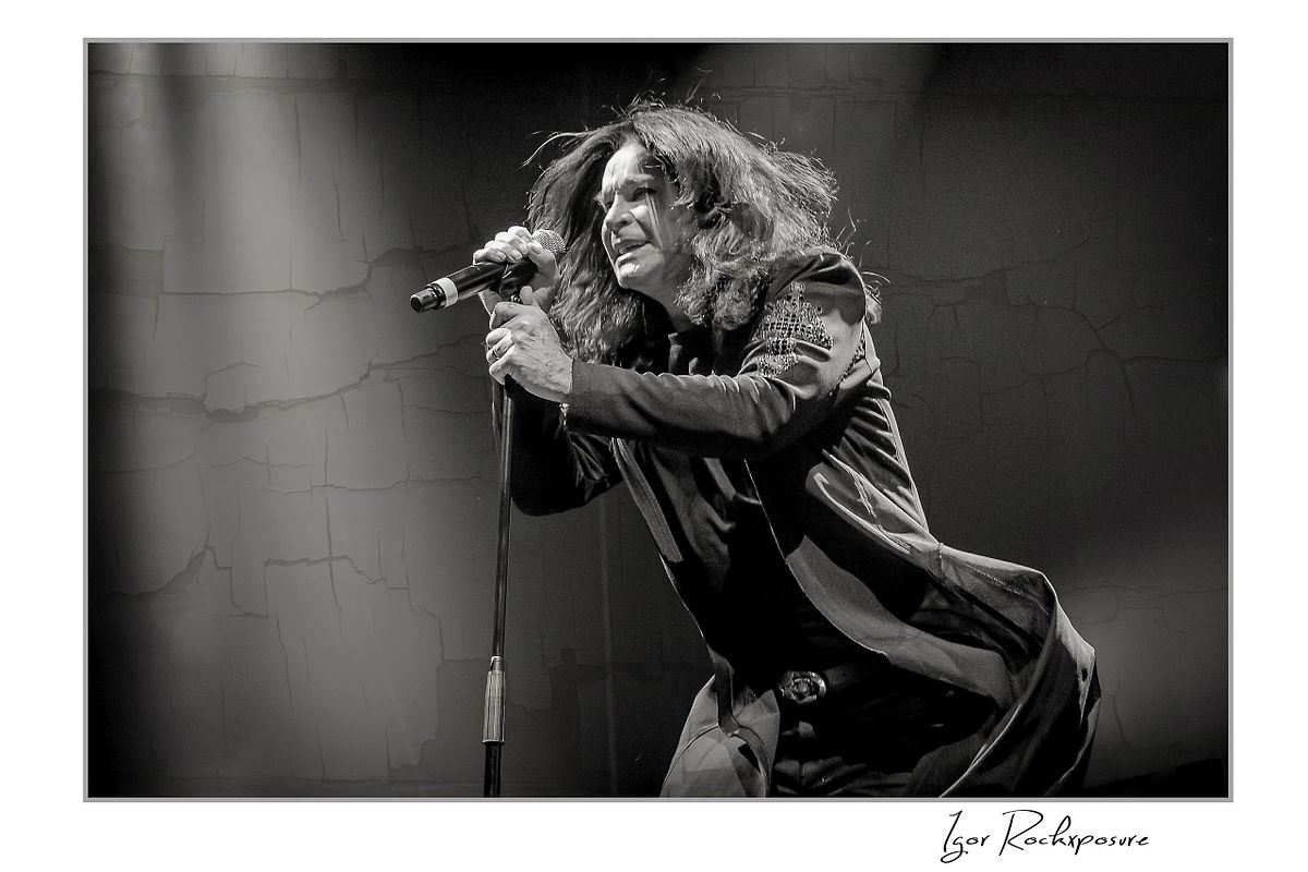 Black and white image of Ozzy Osbourne of Black Sabbath leaning forward singing into a microphone under dramatic stage lighting