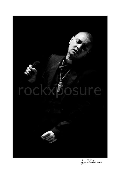 Vertical black and white image of Sinéad O’Connor holding a microphone in a dramatic low-light stage portrait