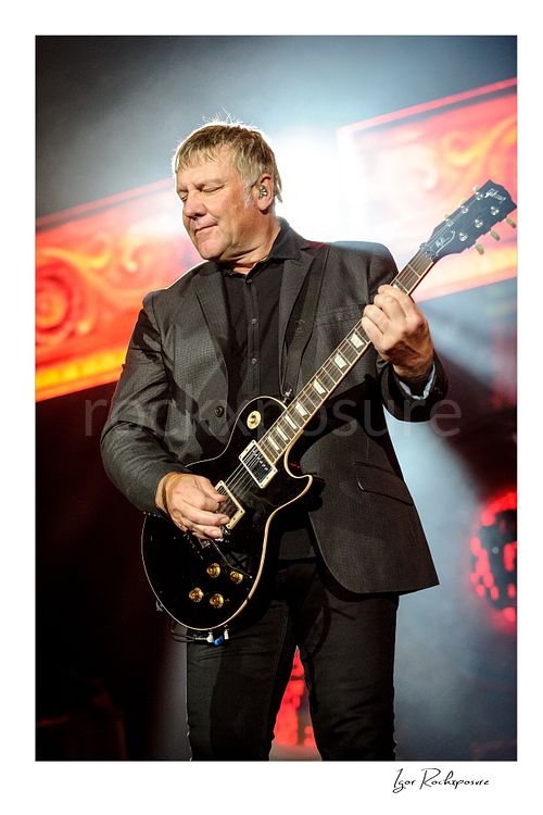 Vertical color image of Alex Lifeson performing with electric guitar during a Rush concert under bright stage light