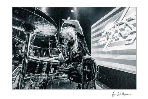 Horizontal black and white image of Robert Sweet of Stryper playing drums with raised drumsticks and flying hair