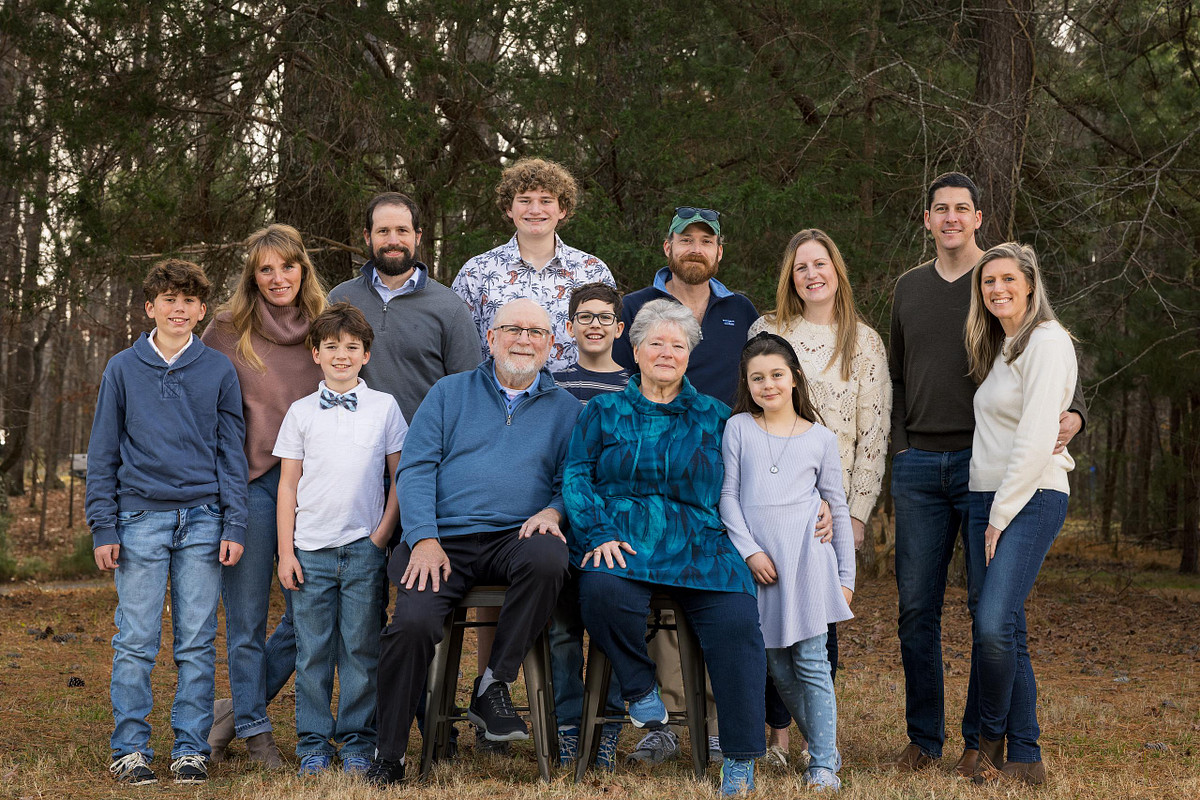 A large extended family of eleven people, including grandparents, three adult siblings, and five children, posing for a group portrait in a wooded area at MLK Park in Carrboro.