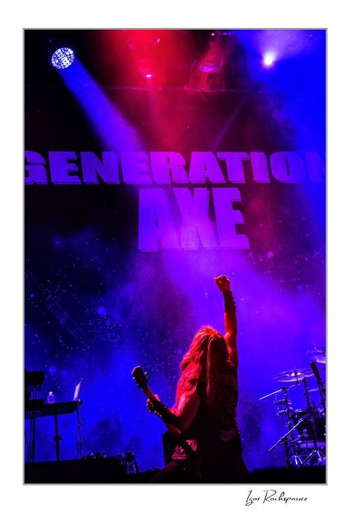 Shot from behind of Zakk Wylde on stage with his guitar, raising his right fist toward a large Generation Axe backdrop under vibrant purple and red lighting.