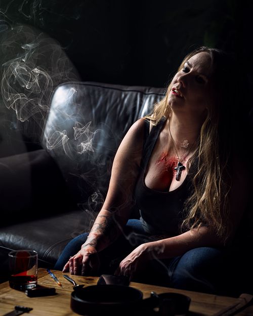 A vampire sitting in the sun, being burned by the cross she wears and glass of blood and a hypodermic needle resting on the table.