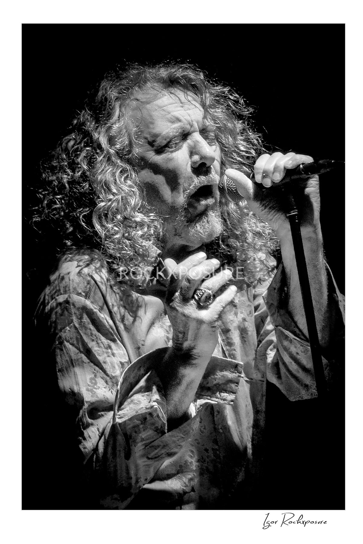 Vertical black and white concert photography of Robert Plant singing into a microphone during a live performance under dramatic stage lighting