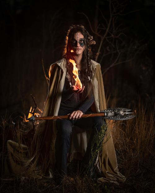 A viking witch wields fire while her battle axe rests on her knees at night.