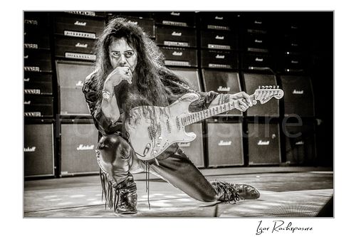 Horizontal black and white image of Yngwie Malmsteen kneeling on stage with a white guitar in a dramatic live performance pose, framed by a wall of Marshall amplifiers behind him