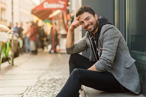 Toni Gojanovic in a gray jacket and striped sitting on the side of the street in Berlin