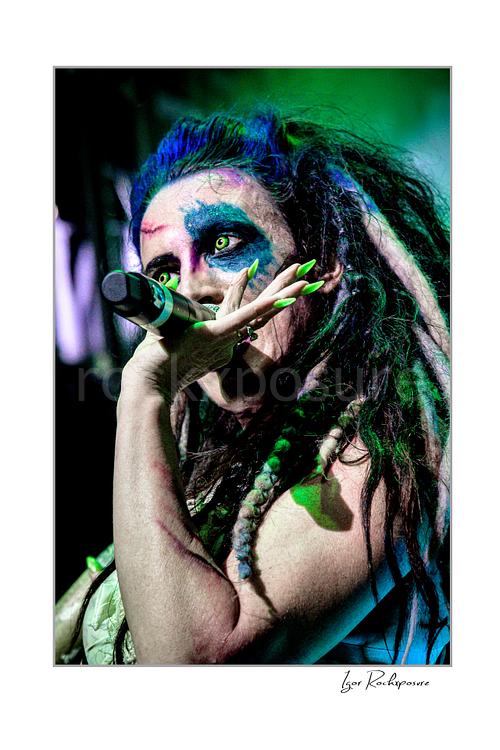 Vertical color image of Calico Cooper of Beasto Blanco singing into a microphone in a tight live performance close-up, with dramatic eye makeup, dreadlocks, and neon green nails under green stage light